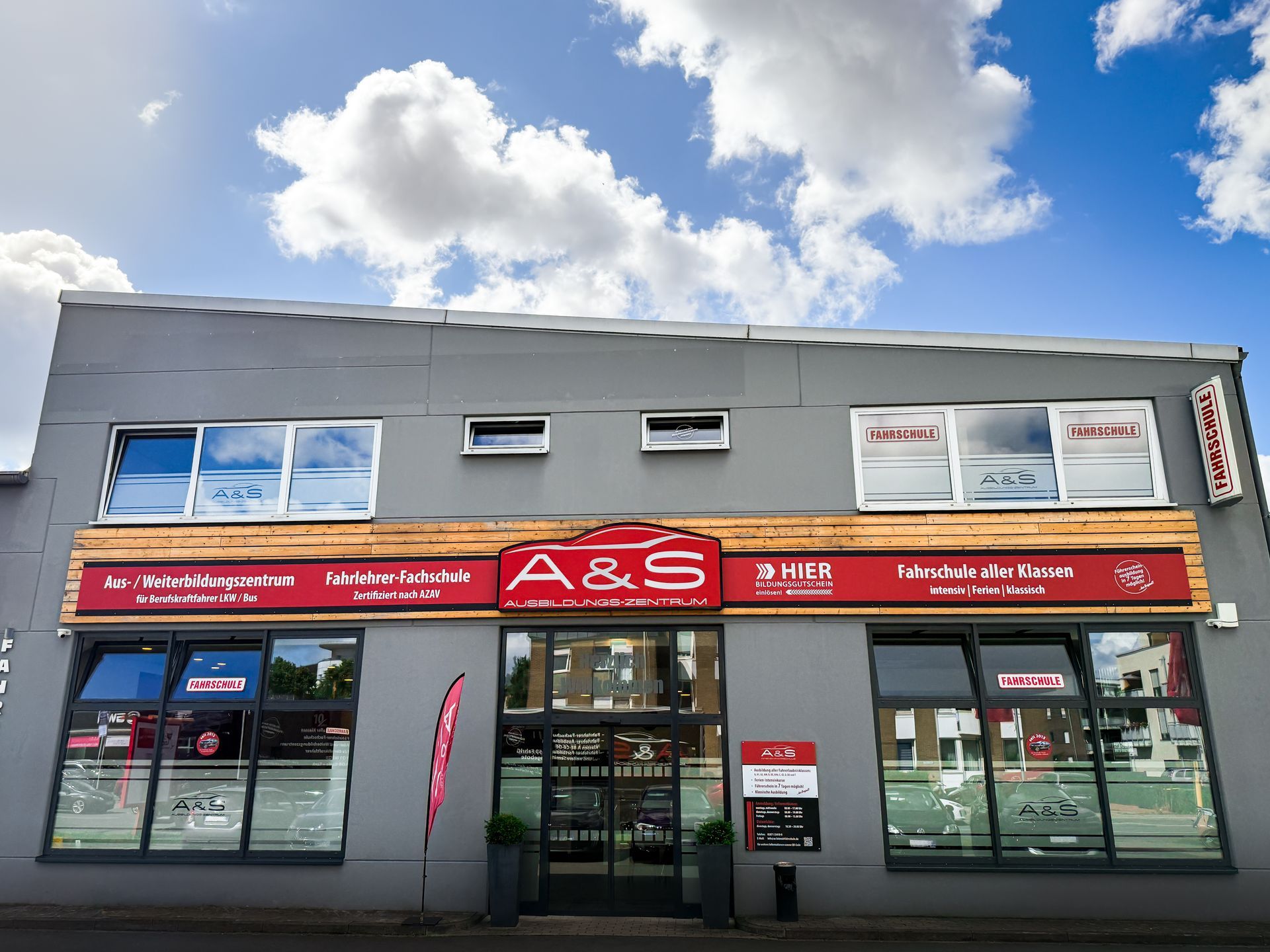 Graues Gebäude mit „A&S“-Schild; Fenster, rote Akzente. Bewölkter blauer Himmel im Hintergrund.