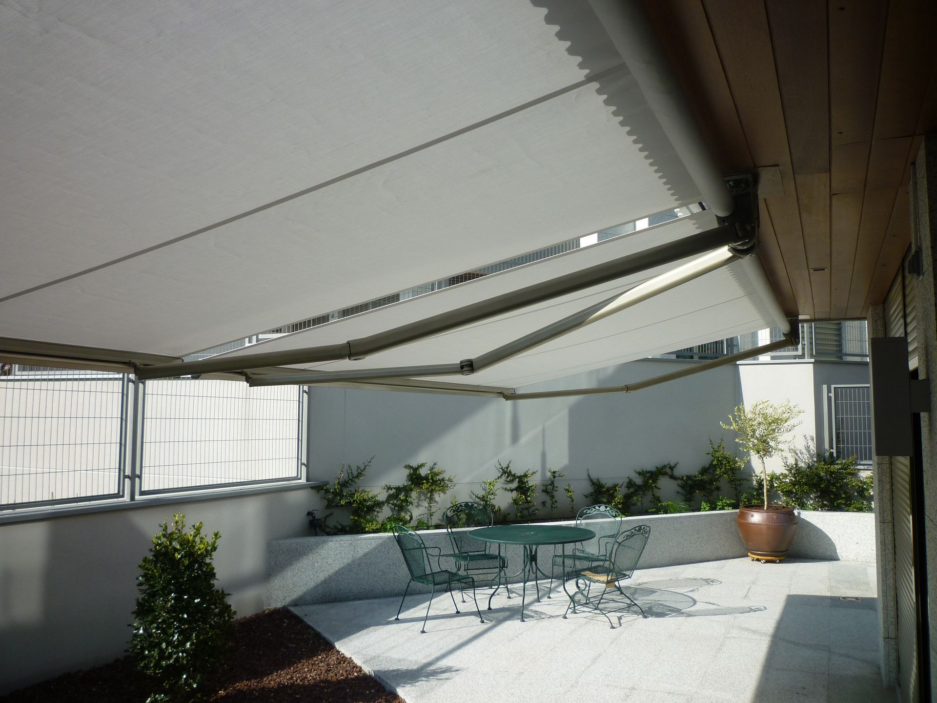 Un toldo retráctil blanco se extiende sobre un patio de piedra que cuenta con una pequeña mesa, sillas y plantas en macetas.