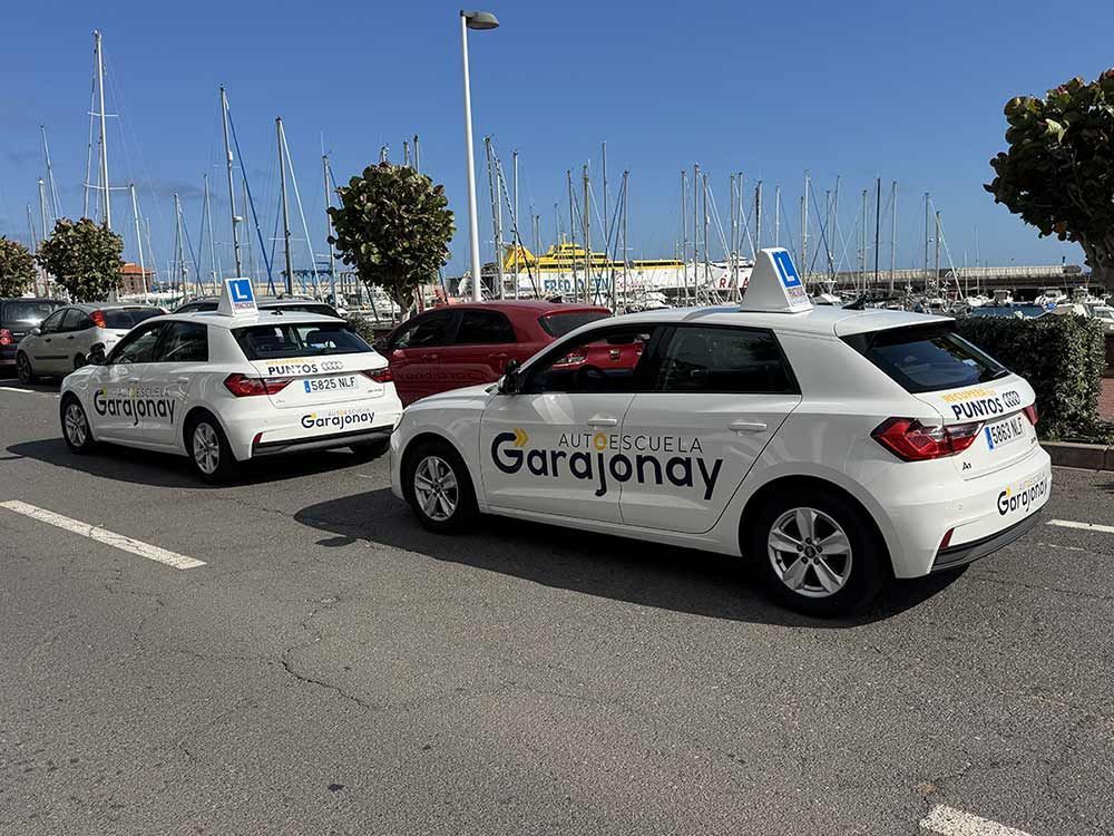 Coches blancos “Garajonay” aparcados junto a un puerto deportivo con mástiles de veleros al fondo bajo un cielo azul despejado.