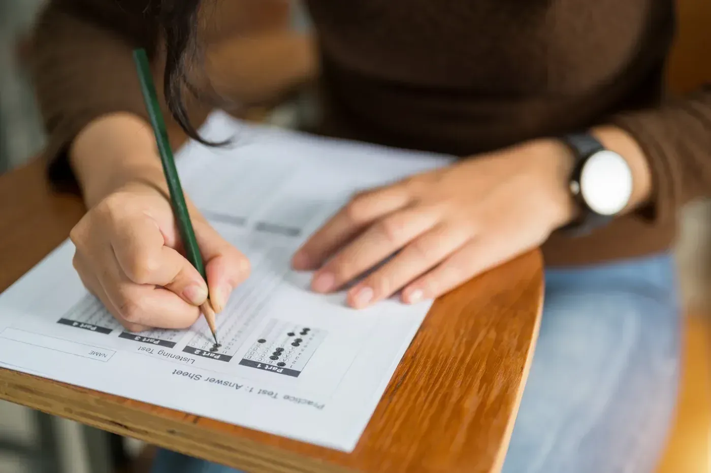 Persona escribiendo en un examen con un lápiz, sentada en un escritorio de madera en un salón de clases.