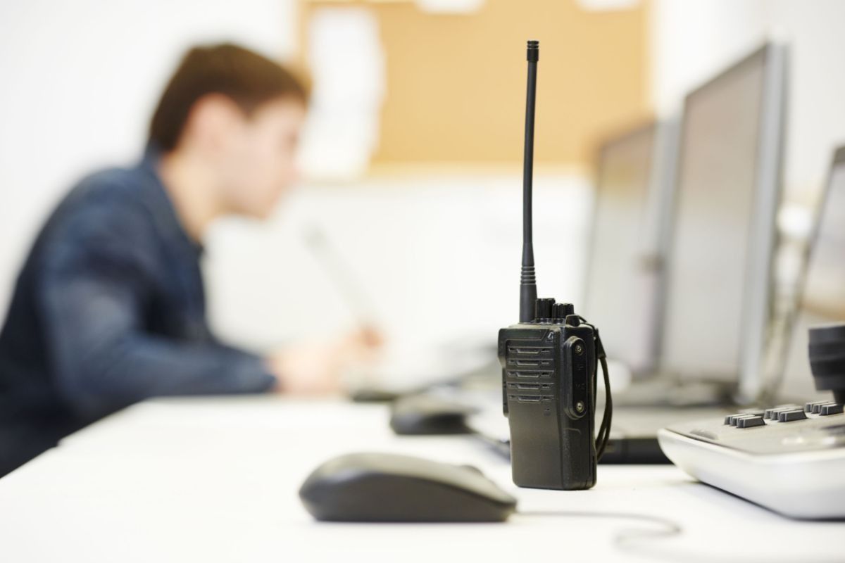 Radio bidireccional sobre un escritorio, con una persona y monitores de computadora en el fondo.