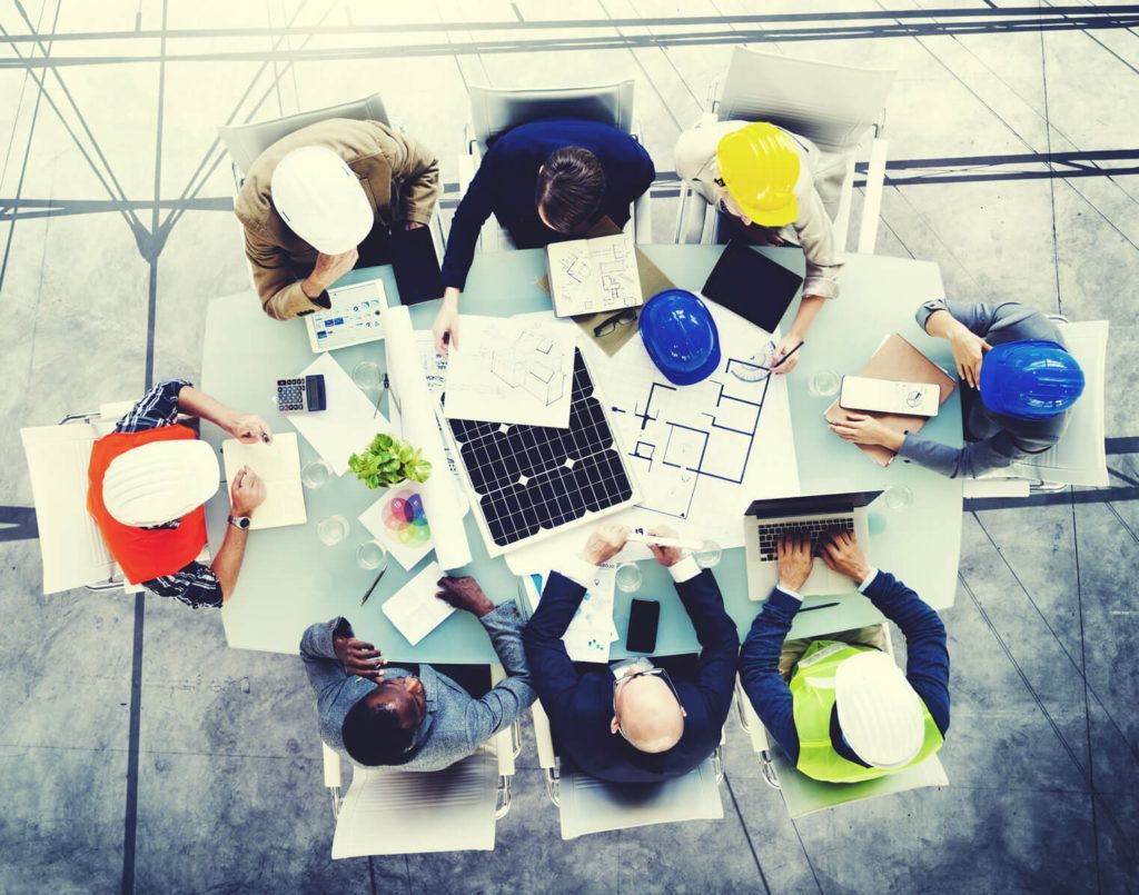 Réunion d'équipe de construction autour d'une table, examinant les plans et les ordinateurs portables ; casques de sécurité de différentes couleurs présents.