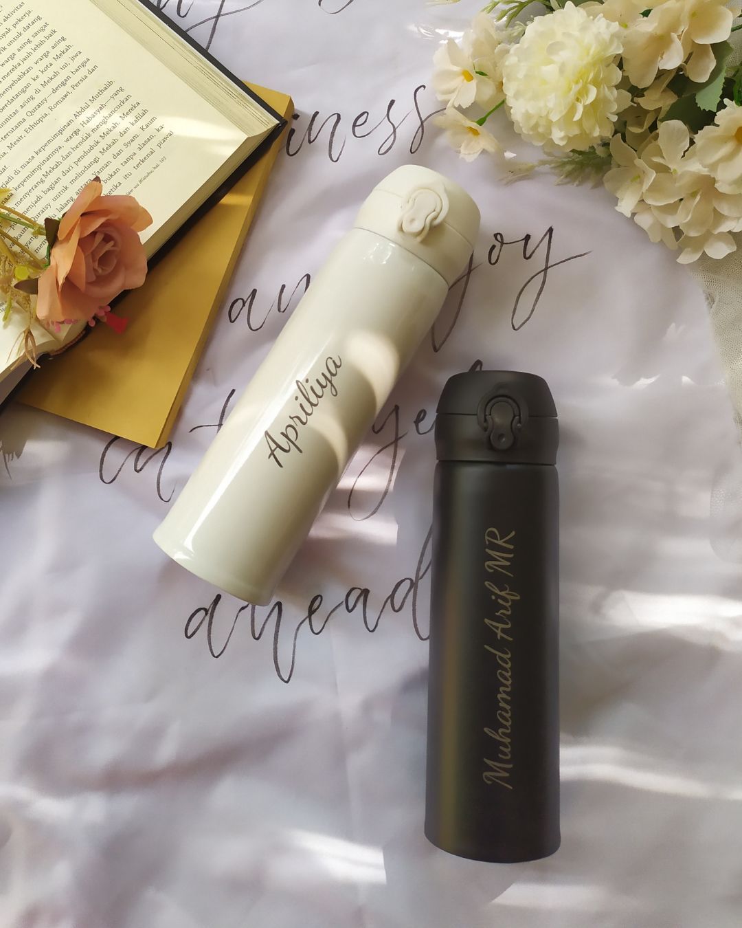 Dos botellas térmicas personalizadas, una blanca y otra negra, sobre una superficie estampada