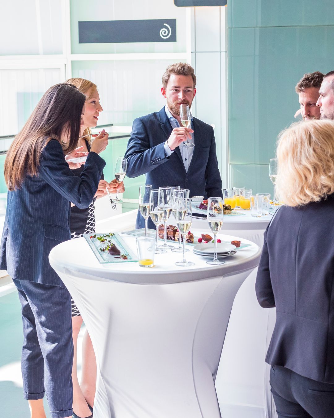 Personas en un evento, bebiendo champán cerca de mesas redondas, dentro de un edificio moderno.
