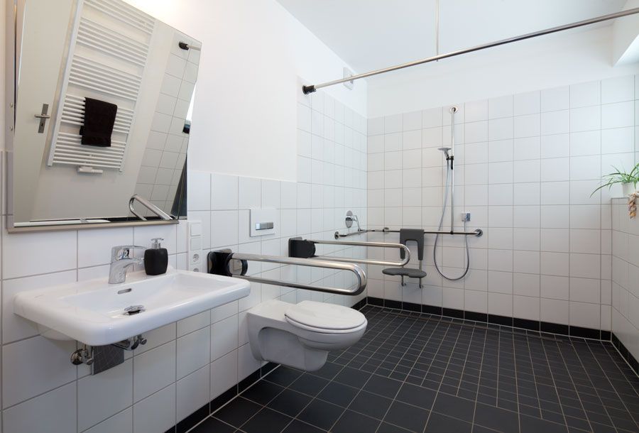 Barrierefreies Badezimmer mit Waschbecken, WC, Haltegriffen, Dusche und schwarzen Bodenfliesen