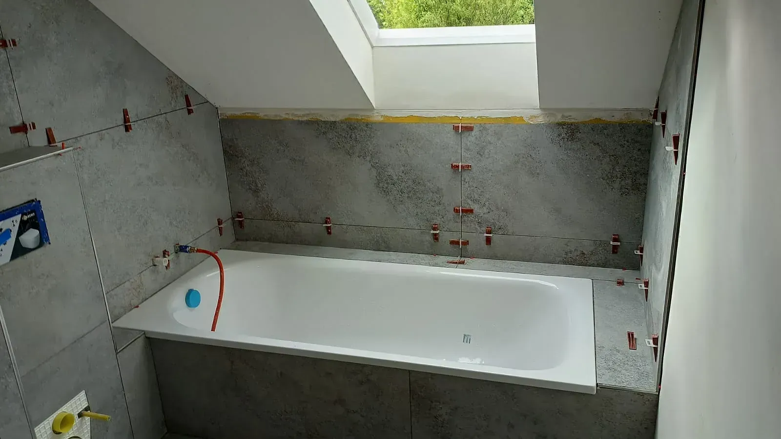 Unfertiges Badezimmer mit eingebauter weißer Badewanne unter einem Oberlicht, umgeben von grau gefliesten Wänden.