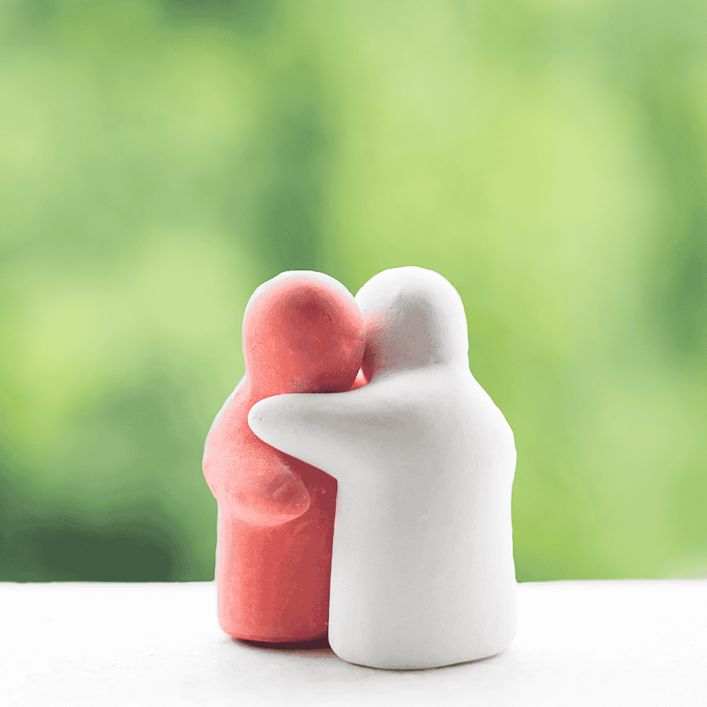 Dos pequeñas figuras de arcilla abrazándose, una roja y otra blanca, con un fondo verde borroso.