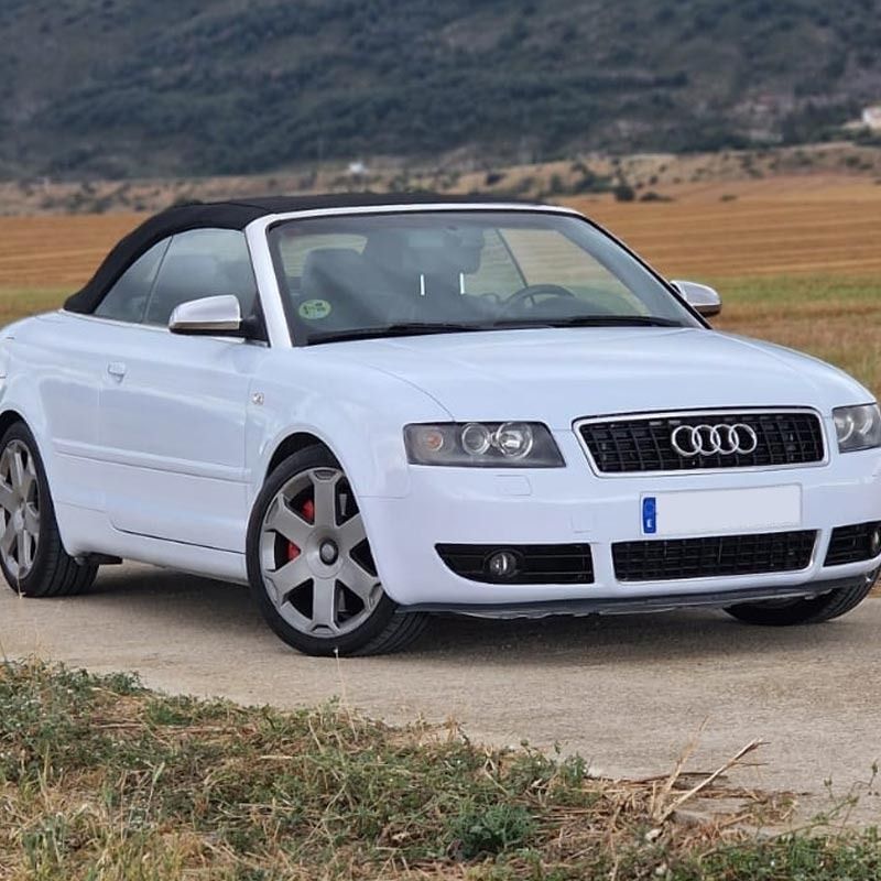 Un Audi blanco descapotable aparcado en un camino de tierra con montañas al fondo. La capota negra está levantada.