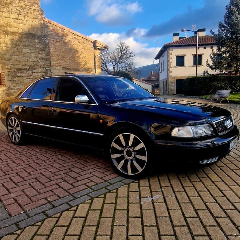 Audi sedán negro estacionado sobre pavimento de ladrillo frente a edificios, día soleado.
