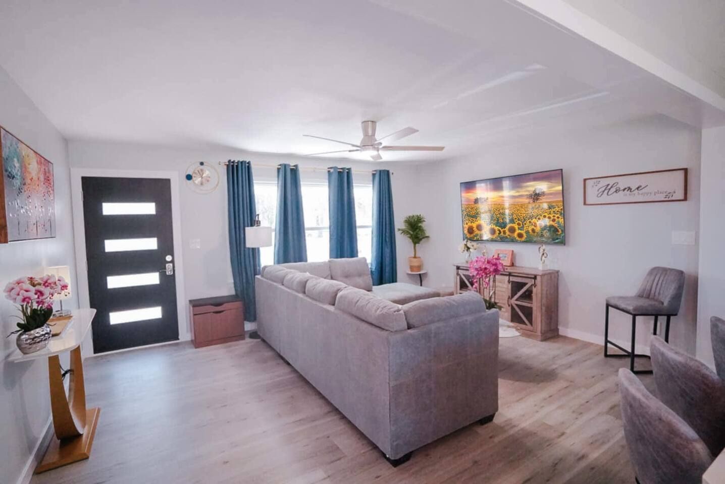 Living room with gray sofa, blue curtains, wooden console, sunflower art, and a black front door.