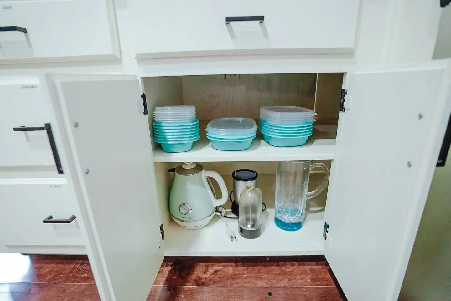 A kitchen cabinet with a kettle , blender , and bowls in it.