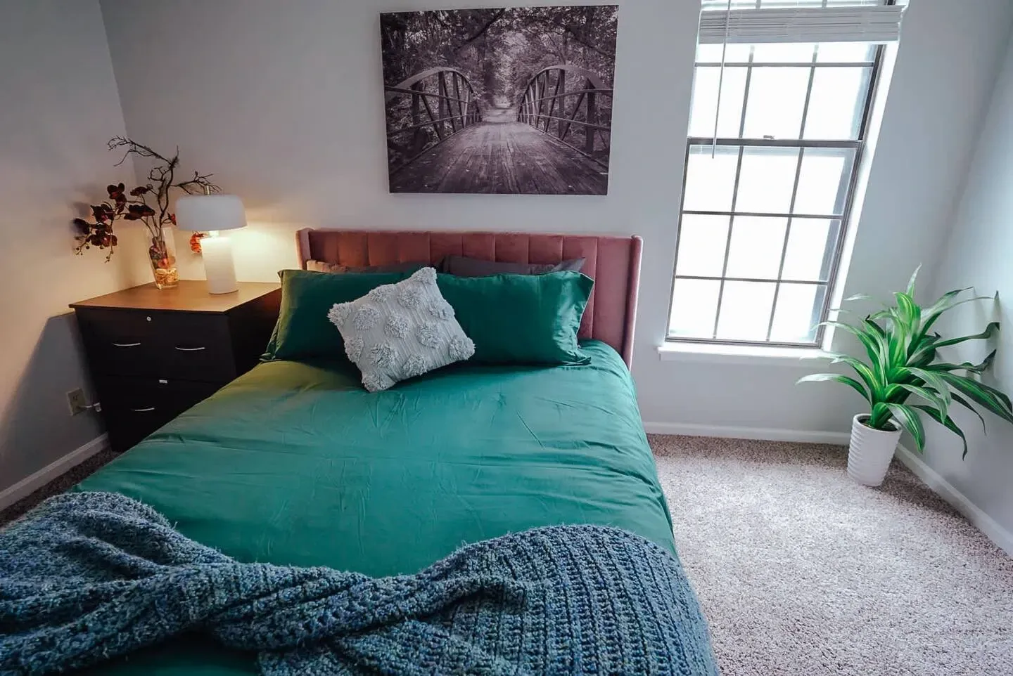 A bedroom with a bed , dresser , lamp and window.