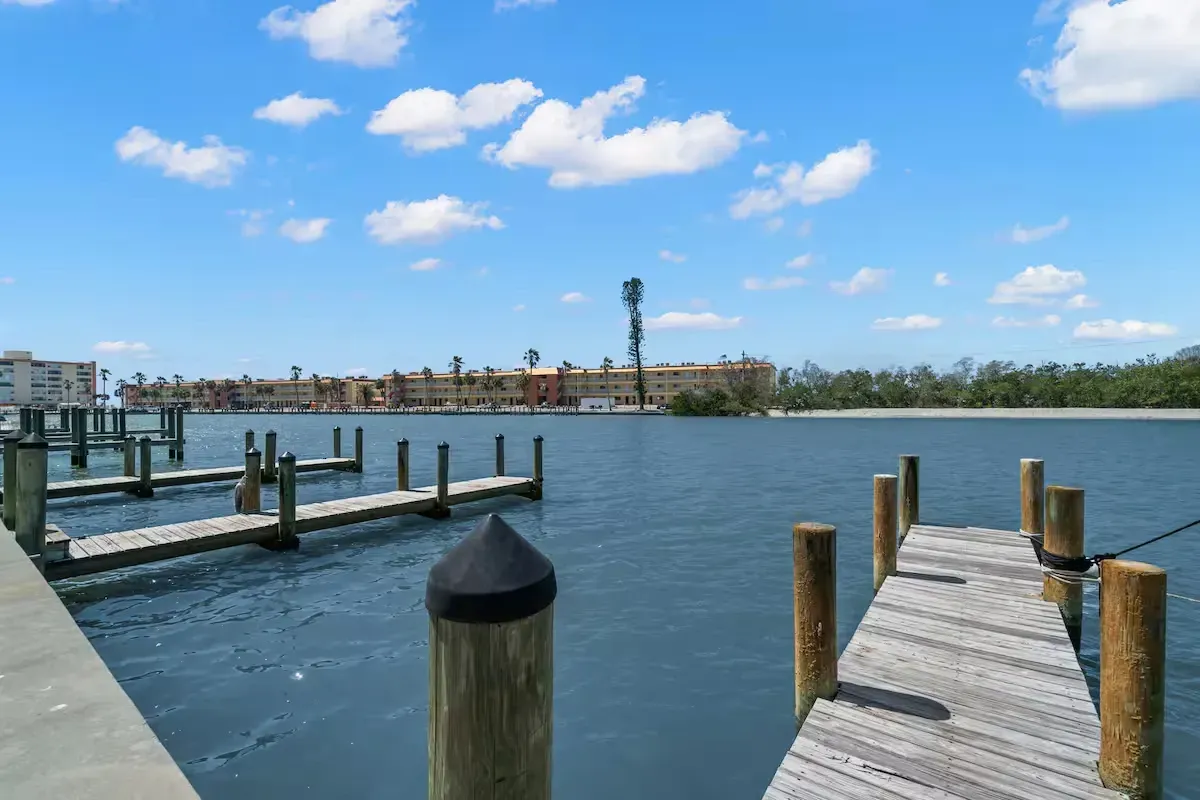 Waterfront view with wooden docks, blue water, buildings in the distance, and a blue sky with fluffy clouds.
