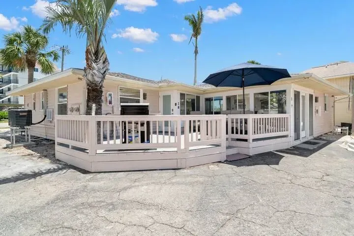 A light pink beach house with a wooden deck and umbrella on a sunny day. Palm trees and a bright blue sky are in the background.