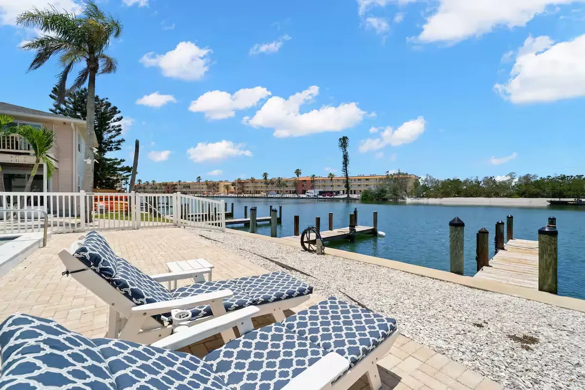 Two lounge chairs on a patio overlooking a canal with a dock, under a blue sky.
