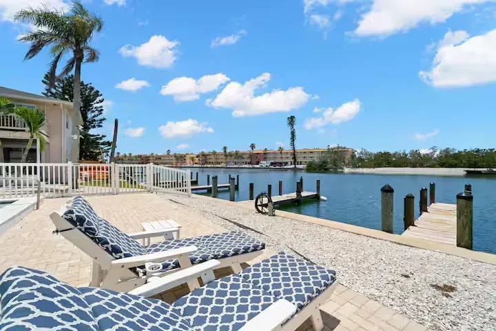 Lounge chairs on a waterfront patio overlooking a marina with blue skies and palm trees.