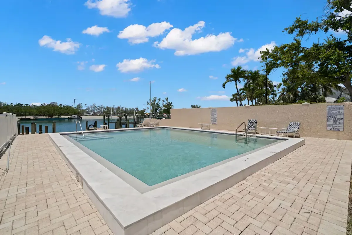 A square swimming pool surrounded by light-colored brick. The sky is blue with scattered clouds, and palm trees are visible.