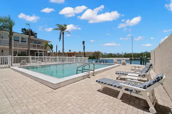 A swimming pool surrounded by a brick patio and white fence, with lounge chairs. Buildings and palm trees in the background under a blue sky.