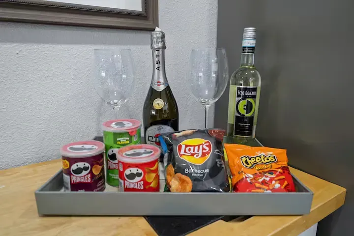 A tray with snacks and drinks, including chips, two bottles of alcohol, and champagne flutes, sits on a table.