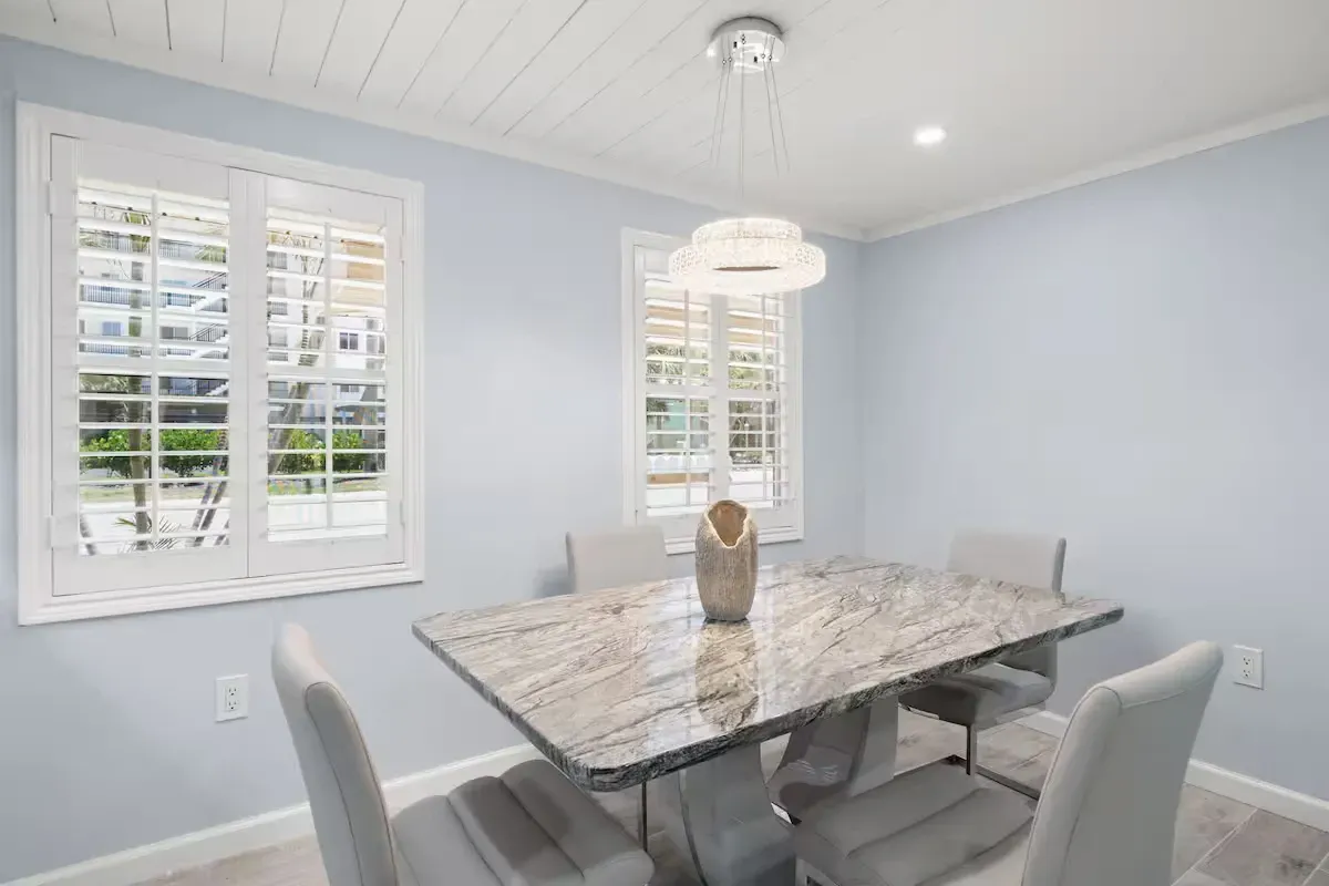 A dining room with a gray marble table, four gray chairs, and two windows with shutters. The walls are light blue.