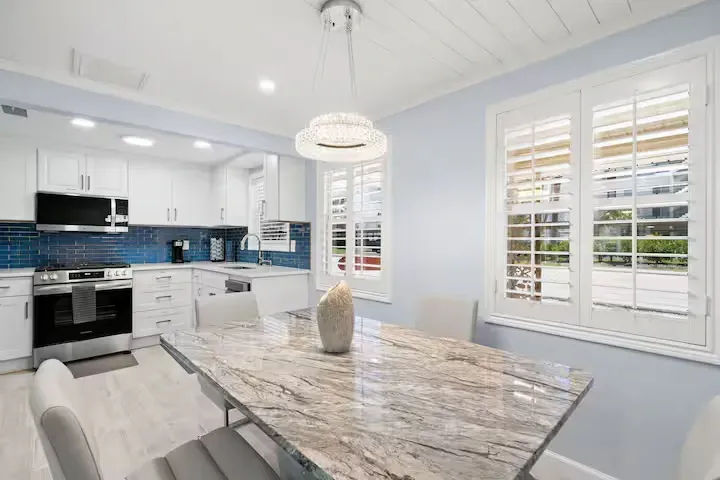 Modern kitchen with white cabinets, blue backsplash, and a marble-topped dining table. White shutters filter sunlight into the dining area.