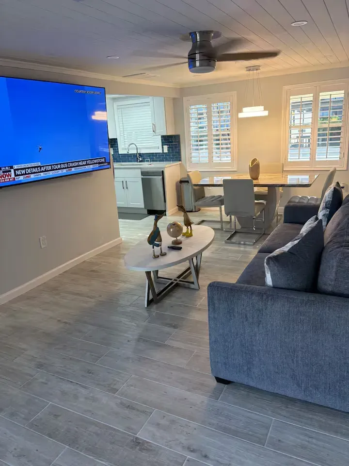 Modern living room with gray tiled floors, a blue sofa, and a wall-mounted TV. Kitchen and dining area visible in the background.