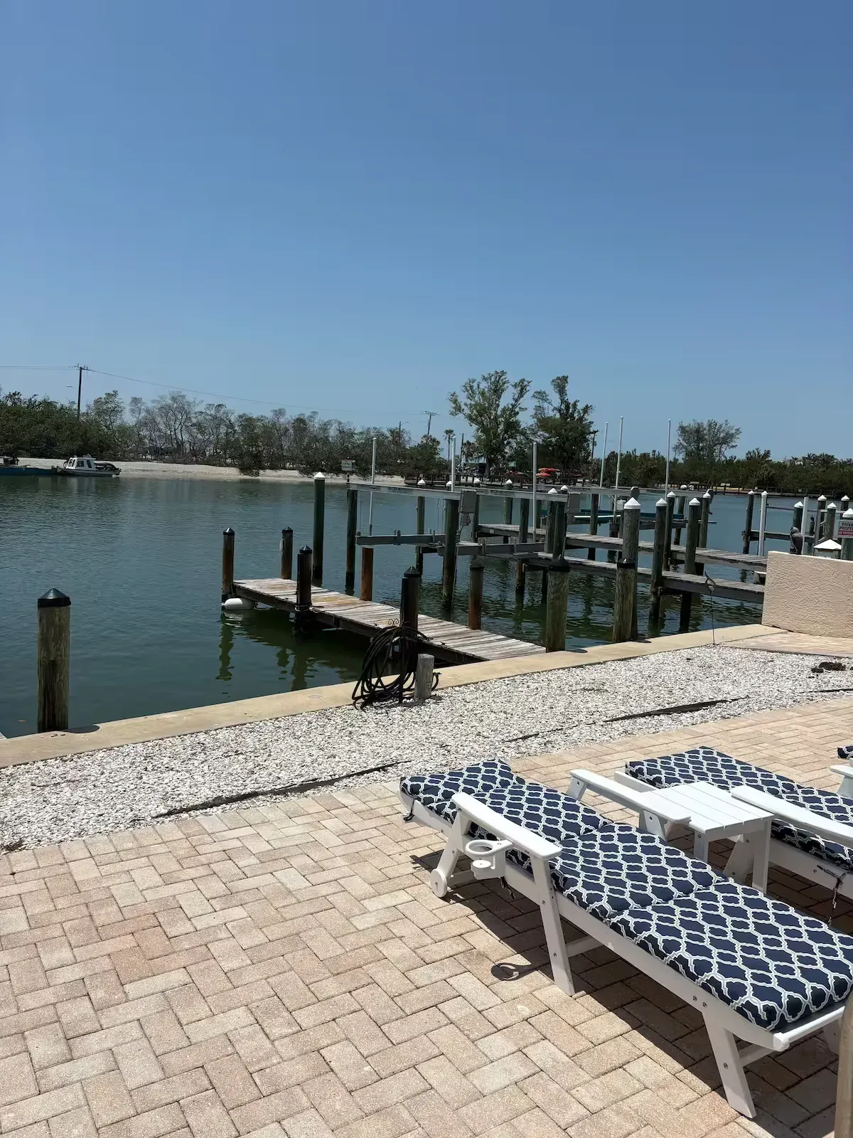 Waterfront scene with docks, calm water, and lounge chairs on a brick patio. Trees and blue sky in the background.