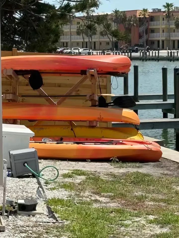 Kayaks stacked on a wooden rack by a body of water, with buildings in the background. The kayaks are orange and yellow.