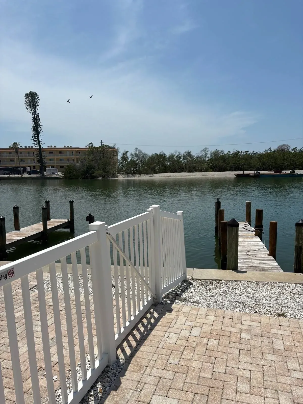 A white picket fence leads to a wooden dock on calm water. Distant shore and buildings are under a blue sky.