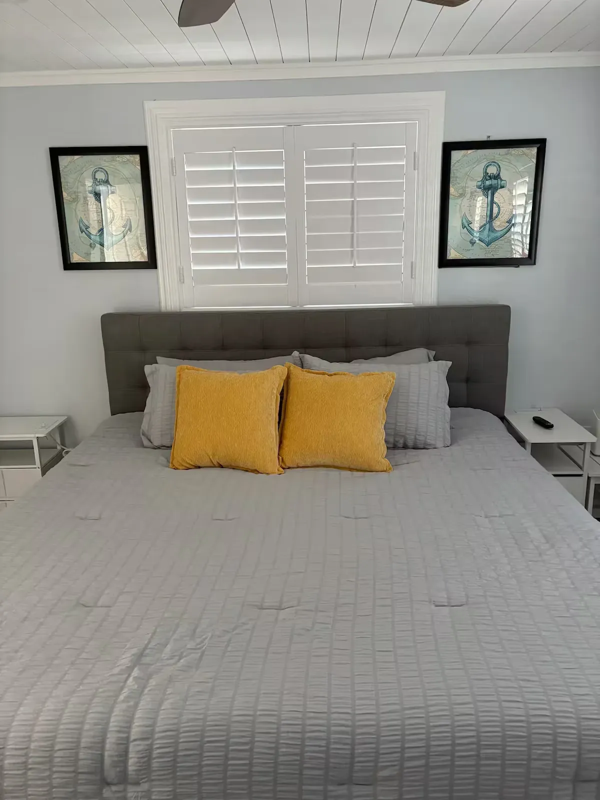 Bedroom with a gray bed, yellow pillows, and two anchor art pieces. White shutters over a window.