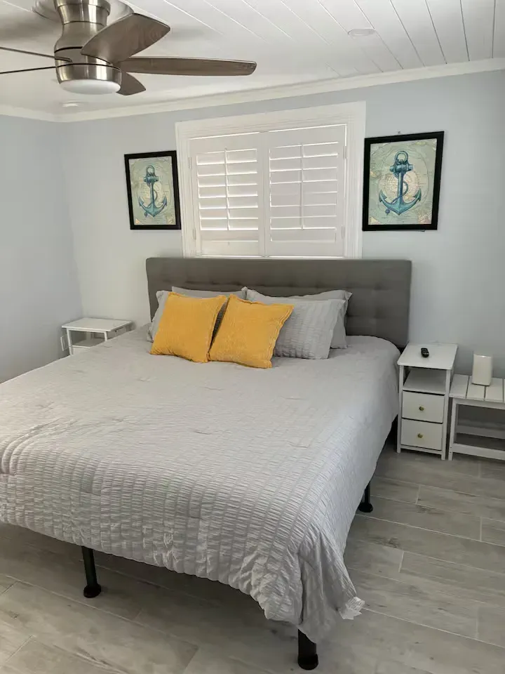 Bedroom with a king-size bed, nightstands, and nautical art. Light blue walls, white shutters, and a ceiling fan.