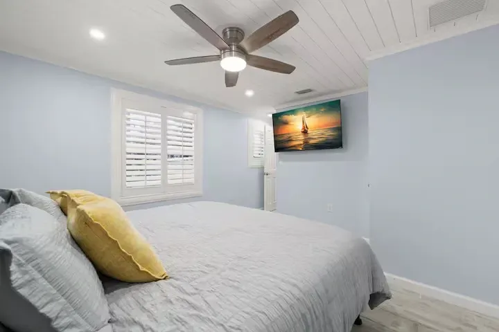 A bedroom with a bed, window with shutters, TV, and ceiling fan; walls are light blue, bed linens are gray.