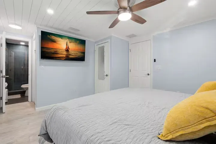 Bedroom with a bed, TV, and bathroom entrance. Light blue walls, a white ceiling, and a sailboat scene on the television.