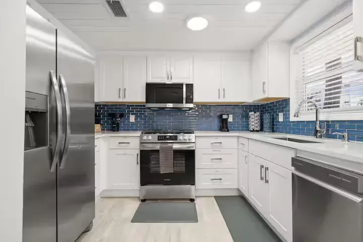 Modern white kitchen with stainless steel appliances, blue tiled backsplash, and light wood-look floors.