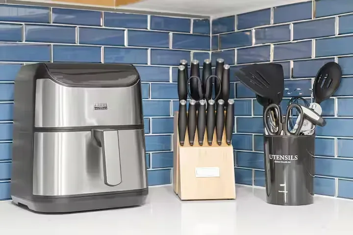 An air fryer, knife block, and utensil holder sit on a countertop against blue tile.