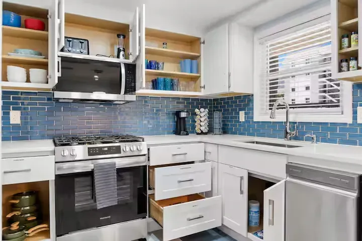 A renovated kitchen with white cabinets, blue backsplash, and stainless steel appliances. The cabinet doors and drawers are open, revealing contents.