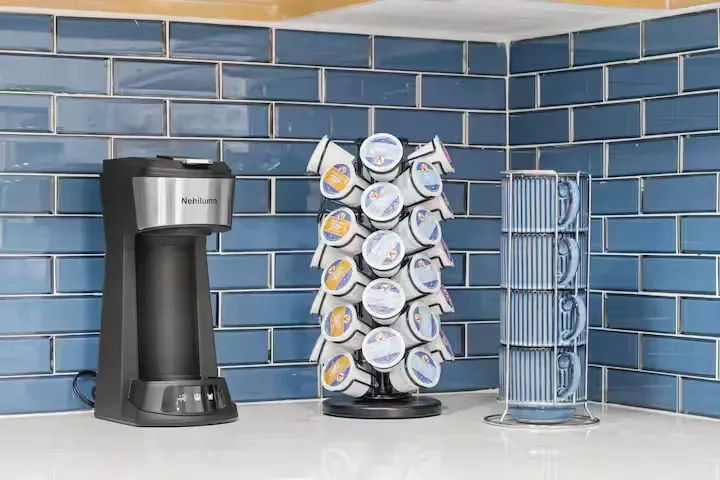 Coffee machine, K-cup carousel, and stacked mugs on a countertop against blue tile backsplash.