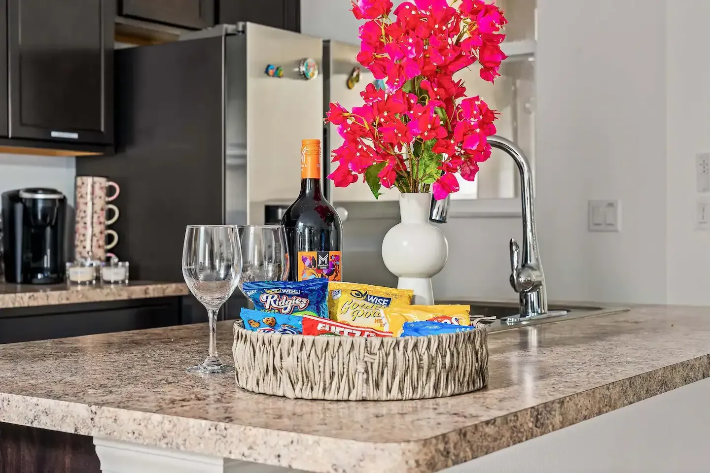 A kitchen counter with a vase of flowers , wine glasses , and snacks.