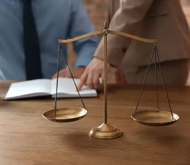 Balanza de la justicia sobre una mesa de madera con dos personas al fondo, una de ellas apuntando a un libro abierto.