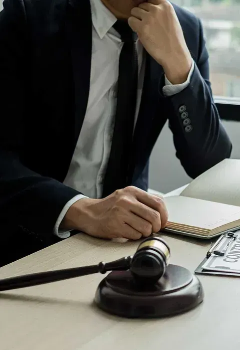 Una persona con traje reflexiona sobre sus pensamientos en un escritorio con un mazo, un cuaderno y documentos legales, probablemente un abogado.