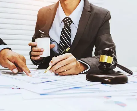 Dos abogados de traje revisan documentos en un escritorio, uno señalando y el otro sosteniendo una taza de café y un bolígrafo cerca de un mazo.