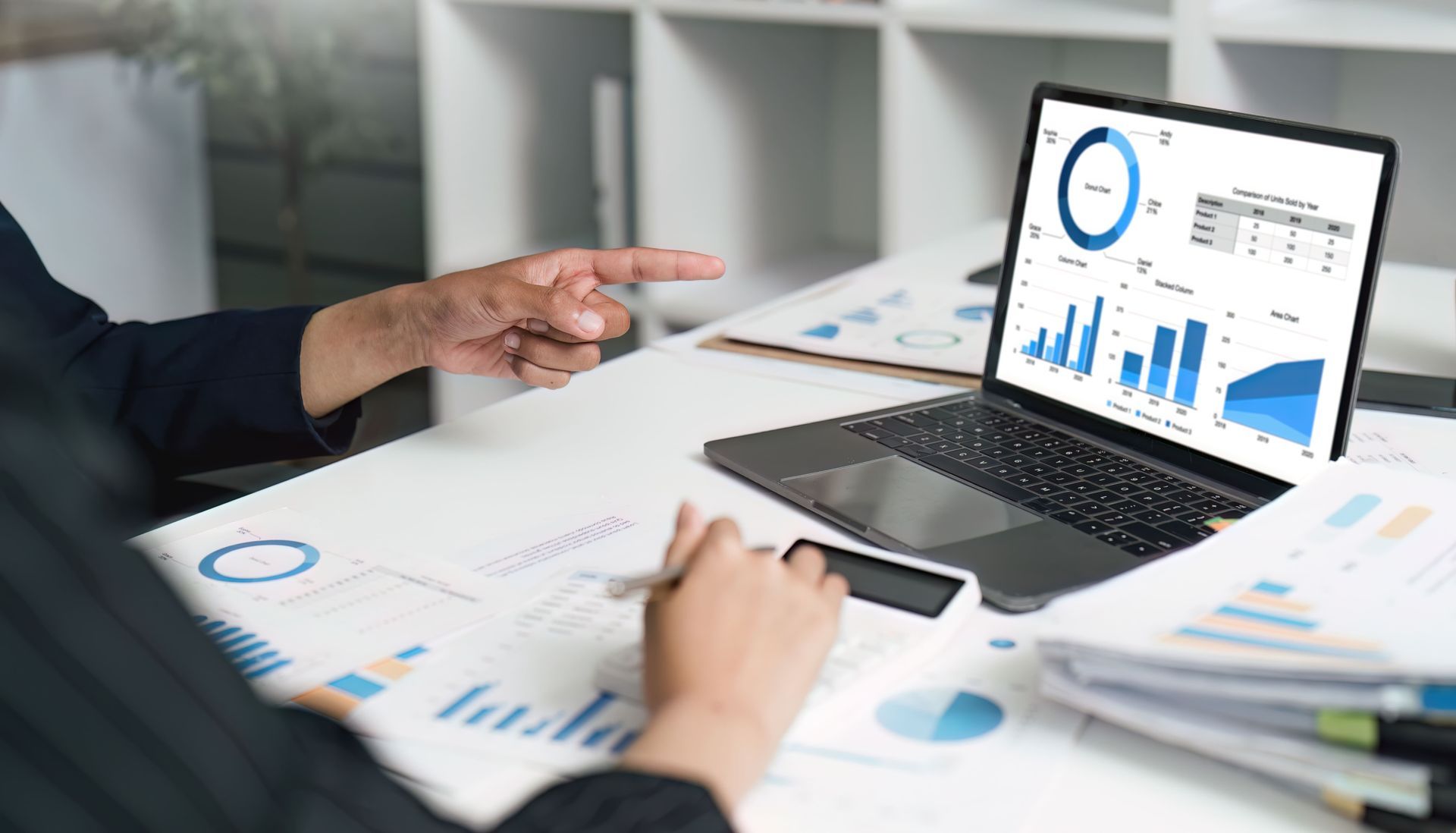 Betriebsprüfung Apotheke: Die Person zeigt auf den Laptop-Bildschirm mit blauen Diagrammen und benutzt dabei einen Taschenrechner, Papiere und Diagramme auf dem Schreibtisch.