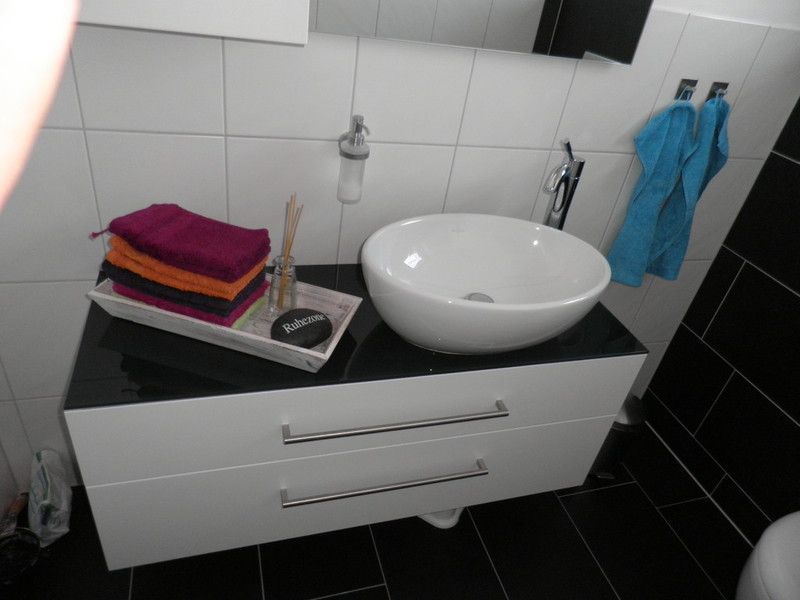 Holzwaschtisch mit schwarzer Oberseite und weißer Front mit zwei Schubladen, die dieselbe Breite wie der Schrank haben. Ein weißes rundes modernes Waschbecken mit verchromter Mischbatterie steht oben rechts auf dem Schrank.