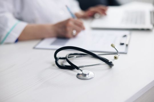 Un estetoscopio está sobre una mesa al lado de un médico que escribe en un portapapeles.