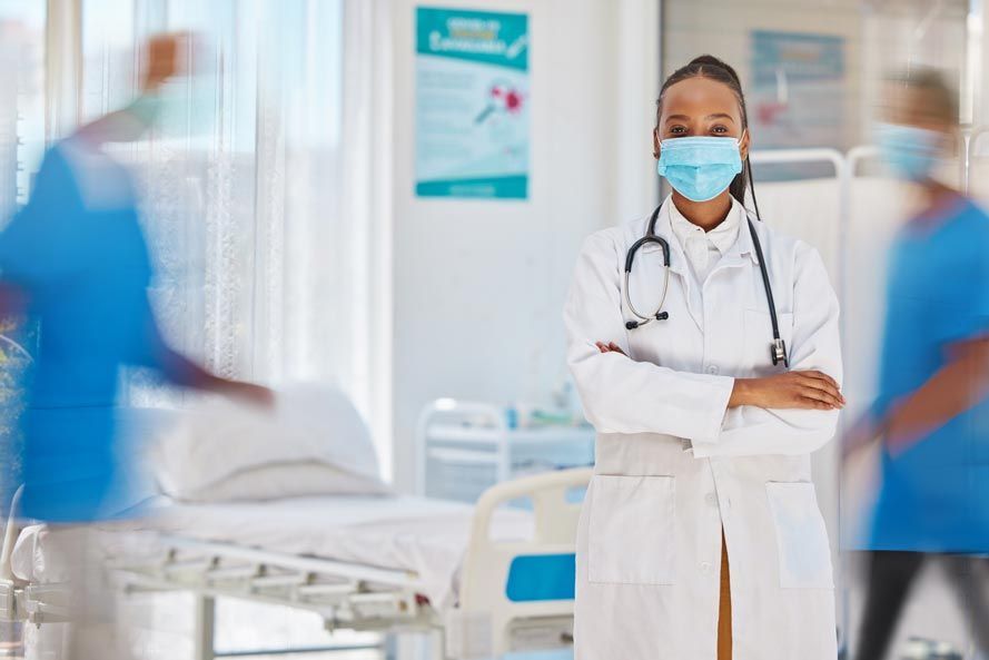 Una doctora que lleva una máscara está de pie en una habitación de hospital con los brazos cruzados.