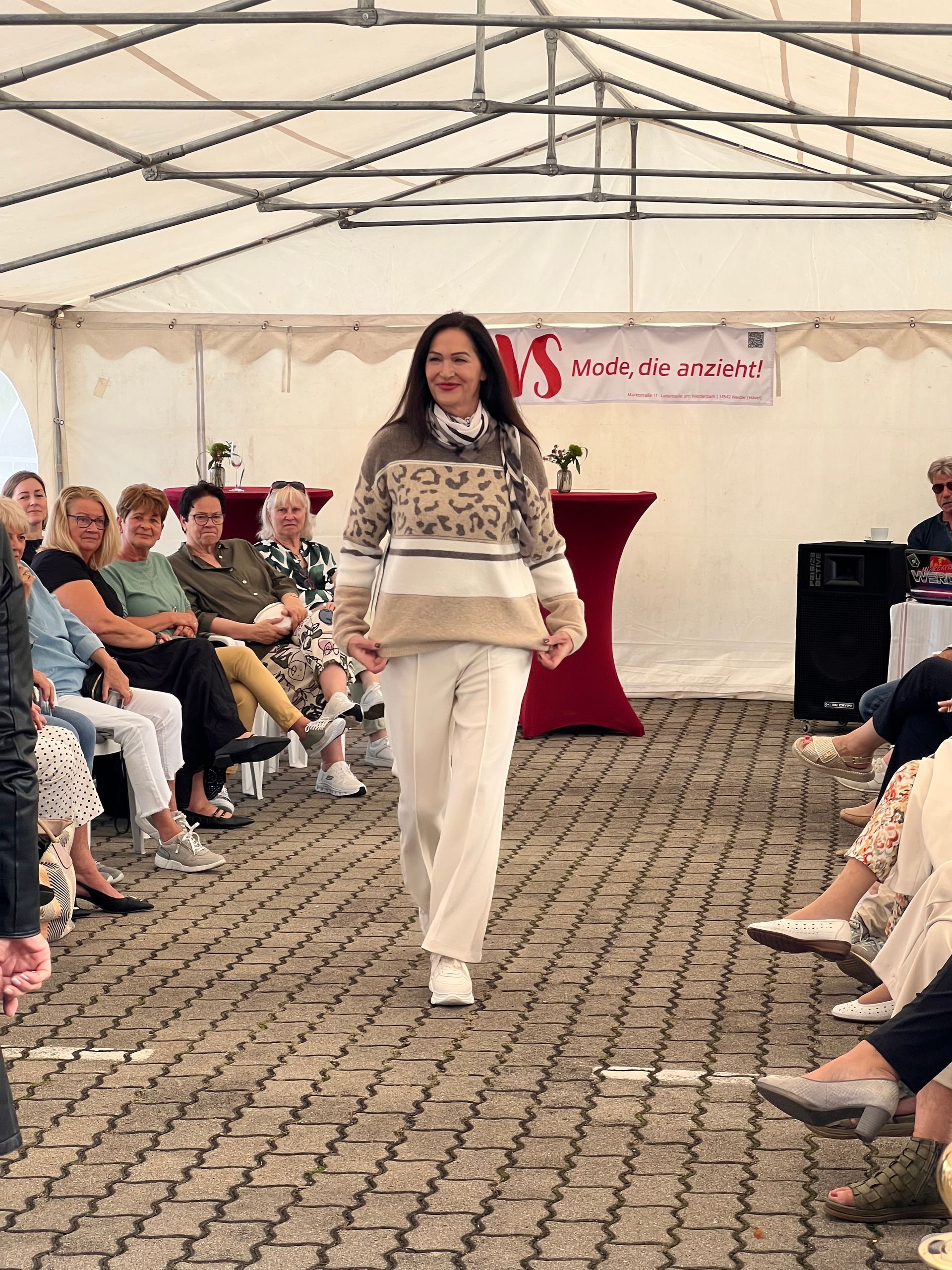 Eine Frau läuft in einem weißen Outfit unter einem Zelt über den Laufsteg, zu beiden Seiten sitzen Zuschauer.