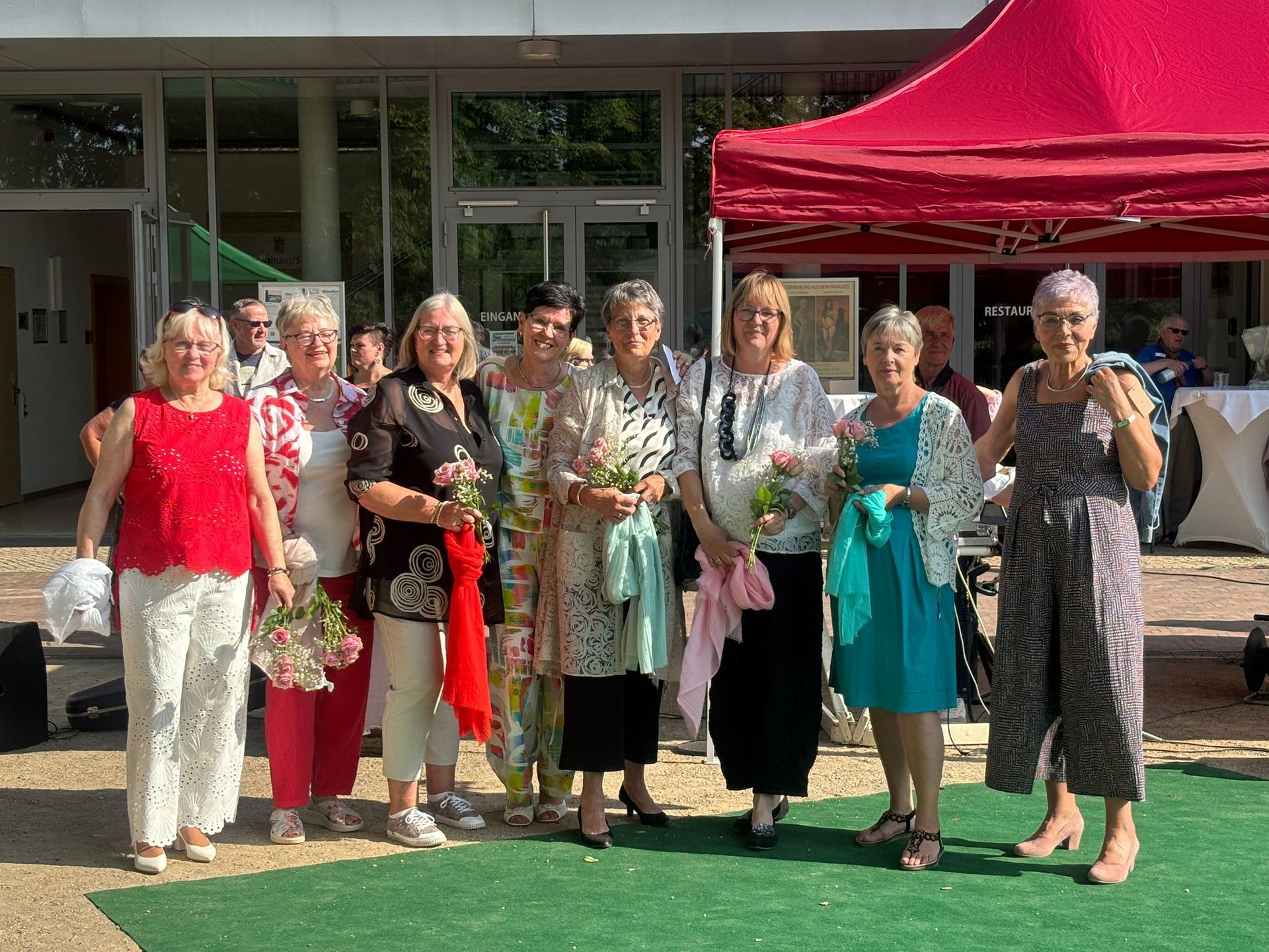 Eine Gruppe von Menschen steht vor einem Gebäude und hält Blumen; in der Nähe befindet sich ein rotes Festzelt.