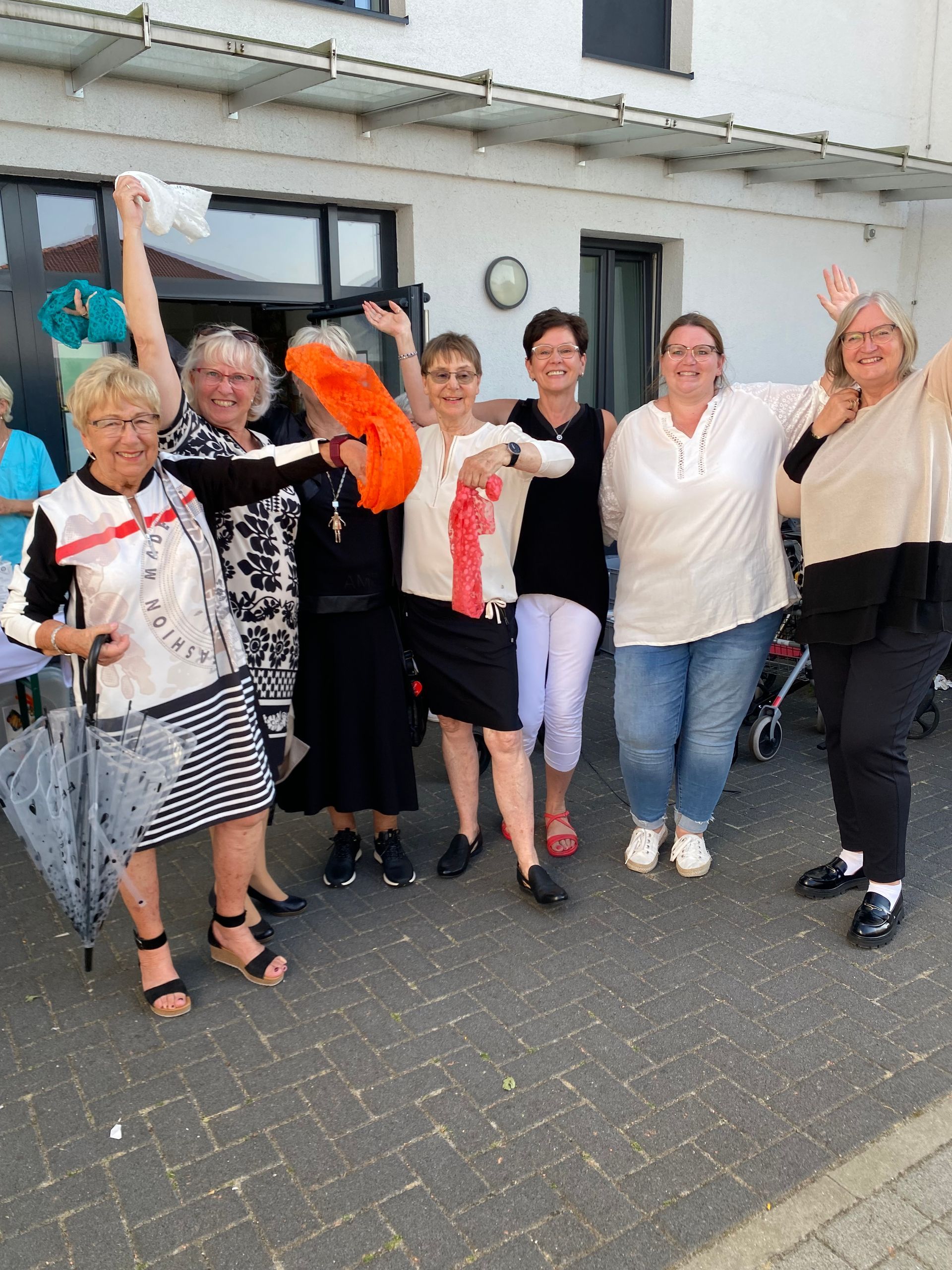 Sieben Personen posieren vor einem Gebäude, lächeln und schwenken bunte Schals bei einer Gruppenfeier.