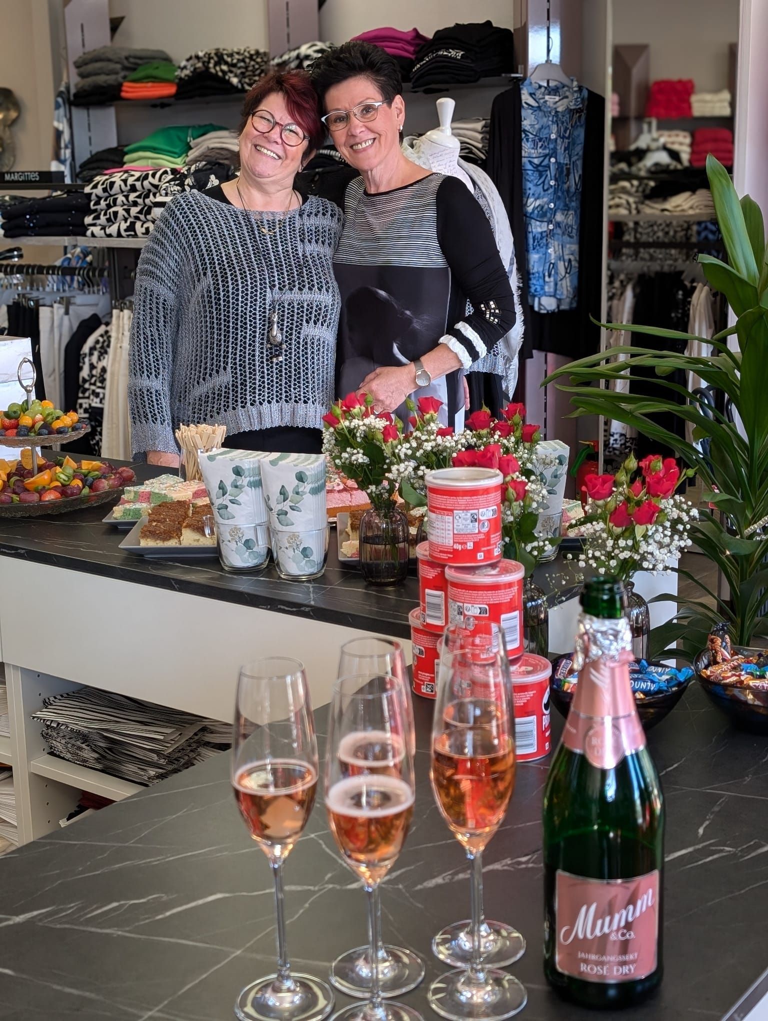 Zwei lächelnde Personen hinter einem blumengeschmückten Tresen, vor dem Weingläser und eine Champagnerflasche stehen.