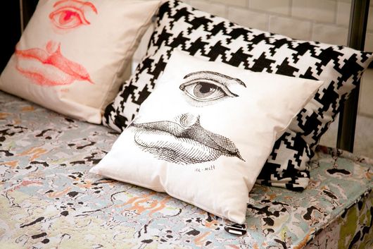 Three decorative pillows on a patterned surface. One pillow has an eye and lips graphic, the other a houndstooth pattern.